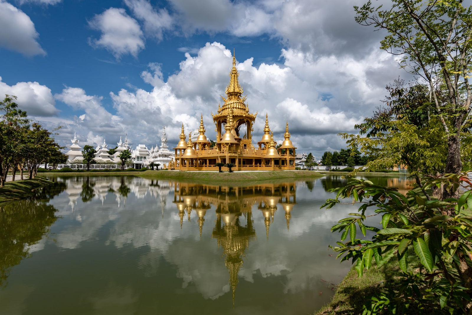 Golden Wat Rong Khun reflected in a serene lake, under a dramatic cloudy sky in Chiang Rai, Thailand.