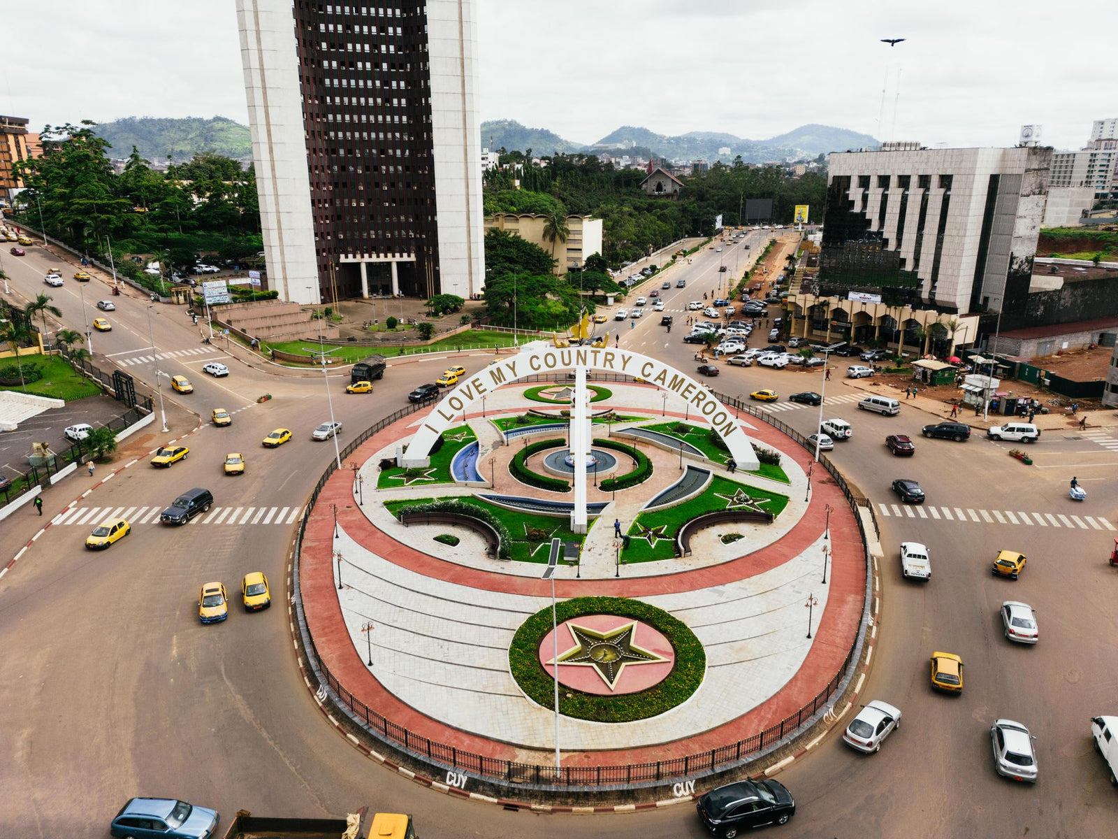 A vibrant aerial view of the 'Love My Country Cameroon' monument in Yaoundé, showcasing urban traffic and cityscape.