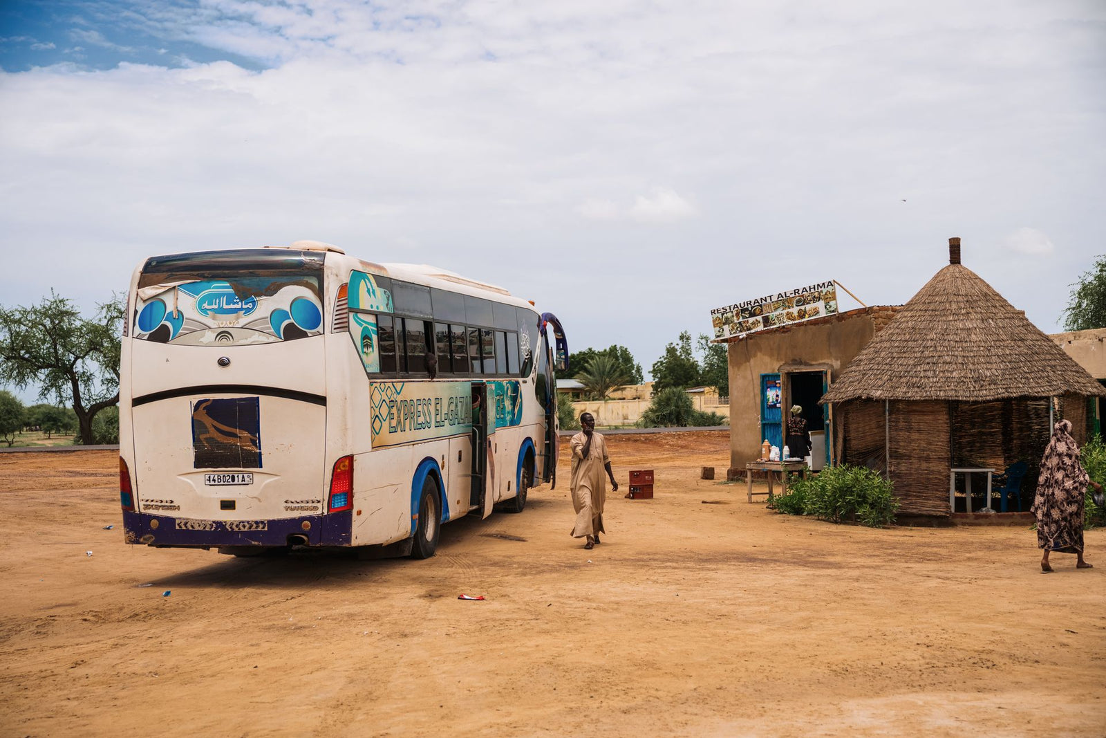 A white bus stops at a rural village with a tribal hut and local restaurant.